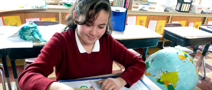 A young student in a red uniform smiles and works on some paperwork at a desk.