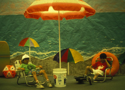 Two students sitting in beach chairs under a beach umbrella in decorated classroom